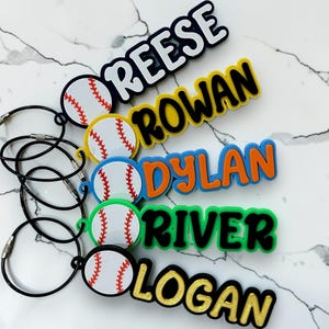 May include: Five baseball-themed keychains with black wire loops. Each features a white baseball with red stitching and a name in raised letters: REESE in white, ROWAN in black, DYLAN in orange, RIVER in black, and LOGAN in gold, all on colored borders.