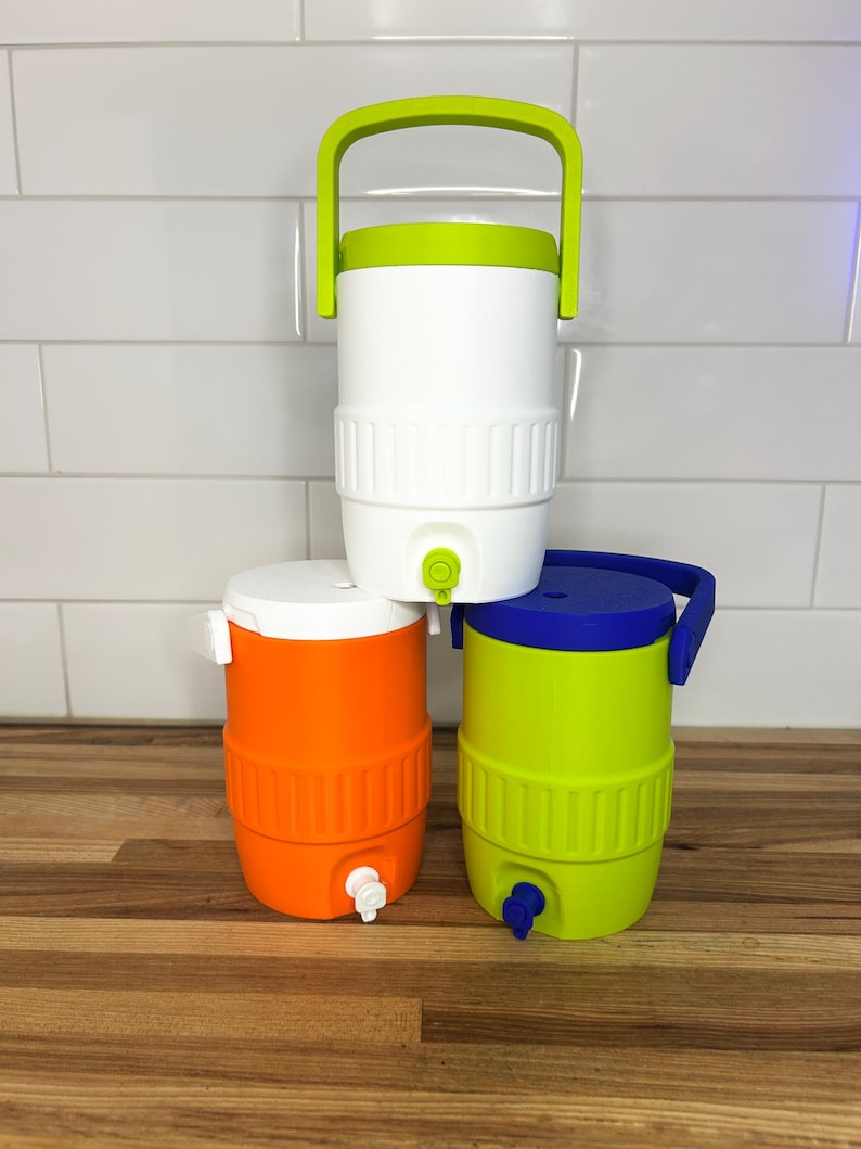 May include: Three colorful water coolers stacked on a wooden surface. The coolers are white, orange, and green, each with a matching lid and handle. The white cooler has a green handle, the orange has a white lid, and the green has a blue lid.