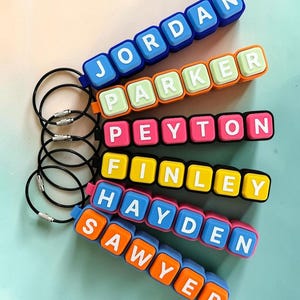May include: Six custom keychains featuring colorful square plastic blocks with white letters spelling the names JORDAN, PARKER, PEYTON, FINLEY, HAYDEN, and SAWYER. Each keychain is attached to a thin black circular metal wire ring with a screw closure.