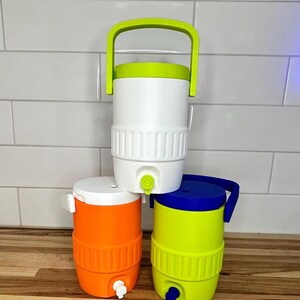 May include: Three colorful water coolers. The top cooler is white with a green handle and lid. The middle cooler is orange with a white lid. The bottom cooler is yellow with a blue handle and lid. Each cooler has a spigot.
