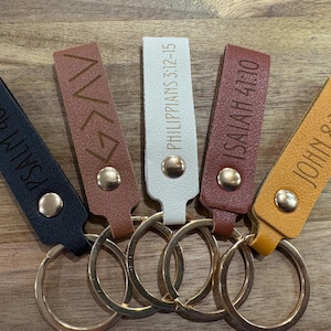 May include: Five leather keychains in black, brown, white, burgundy, and yellow. Each keychain has a gold-colored metal ring and a gold snap. The keychains are engraved with religious text: Psalm 46:10, a symbol, Philippians 3:12-15, Isaiah 41:10, and John 8:12.