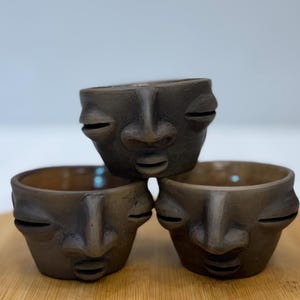 Puede incluir: Tres pequeños maceteros de cerámica marrón oscuro con forma de cara apilados sobre una superficie de madera. Cada macetero tiene un diseño de cara con ojos cerrados y una nariz y boca prominentes. El interior de los maceteros es de color marrón claro.