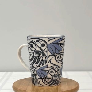 Puede incluir: Una taza de cerámica blanca con asa, con un diseño floral negro y azul. La taza es cilíndrica y se apoya sobre una superficie de madera. El borde de la taza tiene una fina línea azul.