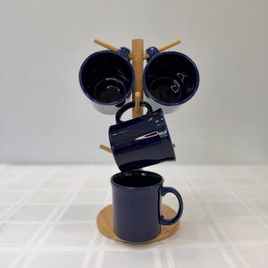 Puede incluir: Un estante para tazas azul marino con cuatro tazas a juego. Las tazas son de color azul oscuro con un acabado brillante y se exhiben en un estante de madera natural. El estante se asienta sobre una base redonda de madera.