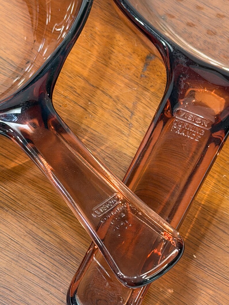 Puede incluir: Dos sartenes de vidrio Vision Corning France con asas. Las sartenes son de color &aacute;mbar transparente. Las asas tienen el logotipo de Vision Corning France. Las sartenes est&aacute;n sobre una superficie de madera.