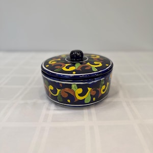 May include: A round, ceramic container with a lid. The container is dark blue with a decorative pattern of yellow, green, and brown swirls. The lid has a small, dark blue knob on top.