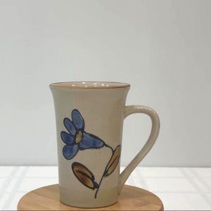 May include: A beige ceramic mug with a blue floral design. The mug has a curved handle and a brown rim. The flower has blue petals and brown leaves. The mug is on a wooden coaster.