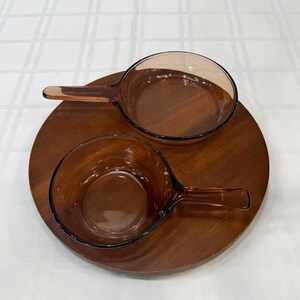 Puede incluir: Dos cuencos de vidrio color &aacute;mbar con asas sobre una bandeja redonda de madera oscura. Los cuencos tienen un dise&ntilde;o cl&aacute;sico y la bandeja tiene un acabado liso y pulido. Este juego es ideal para servir aperitivos o platos peque&ntilde;os.