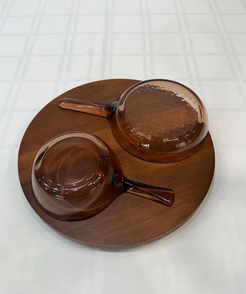 Puede incluir: Dos cuencos de vidrio marr&oacute;n con asas sobre una bandeja redonda de madera. Los cuencos tienen un interior texturizado y son de color &aacute;mbar. La bandeja de madera es de color marr&oacute;n oscuro y tiene un acabado liso. El juego es ideal para servir sopas o salsas.