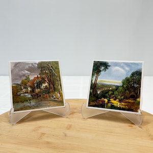 May include: Two square ceramic tiles displayed on clear plastic stands. The left tile depicts a rural landscape with a house, trees, and a small boat on water. The right tile shows a lush green forest scene with a bridge and a dark tunnel entrance.