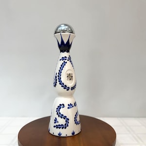 May include: A white ceramic bottle with blue floral patterns and a silver top. The bottle has a bulbous shape with a narrow neck and a wide base. The blue floral pattern is repeated around the bottle.