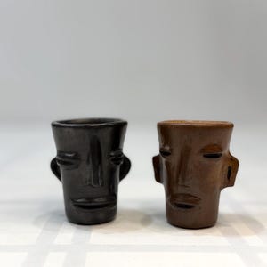 May include: Two small ceramic shot glasses, one black and one brown. Each has a stylized face design with a nose, eyes, and mouth. The glasses have small handles on each side. The glasses are likely handmade.