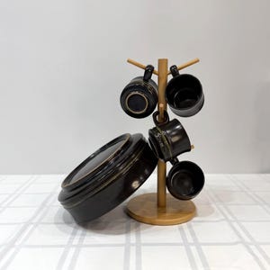 May include: A set of black ceramic mugs and a matching bowl. The mugs are hanging on a wooden mug tree. The bowl is on the table. The mugs and bowl have a dark brown glaze.