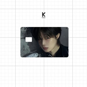 May include: A rectangular card featuring a portrait of a person with dark hair and a black top. The card has a white rectangle and some text on the left side. The letter "K" is above the card.