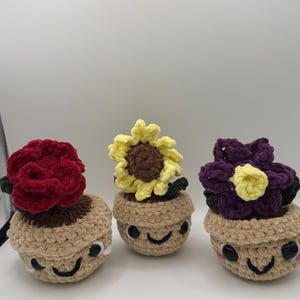 May include: Three handmade crochet flower pot dolls. Each doll has a smiling face and a different flower on top: a red rose, a yellow sunflower, and a purple flower. The flower pots are tan and the flowers have green leaves.