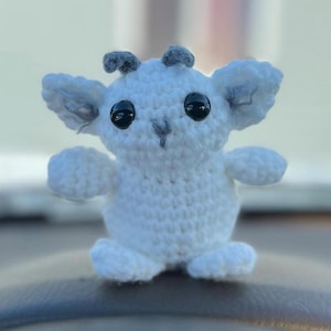 May include: A white crochet creature with large black eyes, grey ears, and small grey horns. The amigurumi figure has a rounded body, small arms, and feet. The plush toy is a whimsical, soft sculpture.
