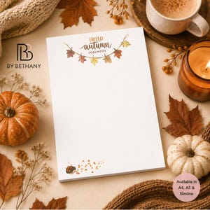 May include: A white notepad featuring a decorative garland of autumn leaves at the top with the text Hello autumn LISA'S NOTES and a small illustration of a hedgehog at the bottom. The notepad is surrounded by pumpkins and fall foliage.