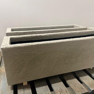 Peut inclure: Trois jardinières rectangulaires de couleur claire avec une surface texturée. Les jardinières ont un intérieur sombre et sont empilées sur une palette en bois. Convient pour l'extérieur ou l'intérieur.