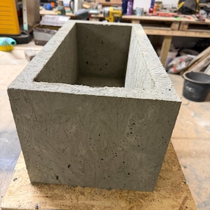 Può includere: Una fioriera rettangolare in cemento con una superficie ruvida e grigia. La fioriera è aperta nella parte superiore e poggia su una base di legno. La scatola è progettata per l'uso all'aperto ed è adatta per piantare fiori o piccole piante.