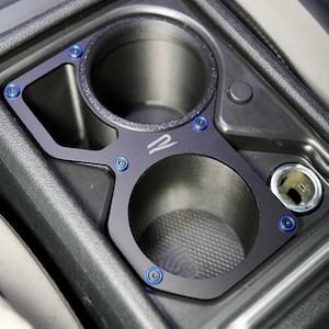 以下が含まれることがあります： 車両のセンターコンソール用ブラックのデュアルカップホルダーインサート。マット仕上げで、白いロゴとフレームを固定する青い金属製ボルトが特徴です。このユニットは、電源ソケットの隣のコンソールスペースに収まるように設計されています。