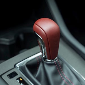 May include: A car gear shift knob featuring a deep red leather-like top section with a smooth texture, accented by a metallic silver base. The black gear shift boot is finished with red stitching, and the gear indicator panel displays P, R, N, D, M.
