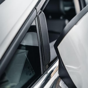 Puede incluir: Un marco de puerta de coche blanco con una moldura de pilar vertical negra, una carcasa de espejo lateral y un cristal de ventana con marcas visibles. El panel exterior es blanco, contrastando con las juntas de goma negras.