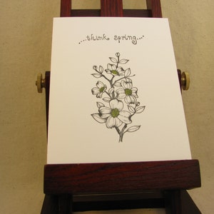 May include: A white card with a black and green floral illustration and the words "...think spring..." in black script. The card is displayed on a small wooden easel with brass hardware.