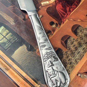 Peut inclure: Un couteau à beurre argenté avec un manche détaillé représentant un ours tenant un poisson, des montagnes et des arbres. La lame du couteau est lisse et brillante. Le manche est sculpté en relief.