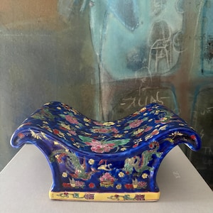 May include: A ceramic headrest featuring a deep blue glaze decorated with colorful painted dragons, phoenixes, and floral motifs. The curved, saddle-shaped top rests on a rectangular base with a yellow border and intricate floral patterns.