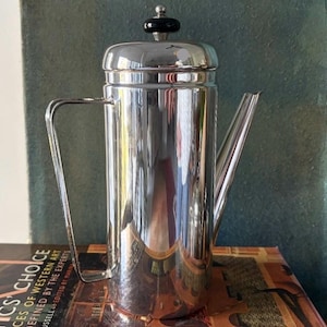 Puede incluir: Una cafetera alta de acero inoxidable pulido con asa y boquilla. La cafetera tiene una tapa con un pomo negro. Está sobre un libro con el texto "CS CHOICE PIECES OF WESTERN ART AND DEFINED BY THE EXPERTS".