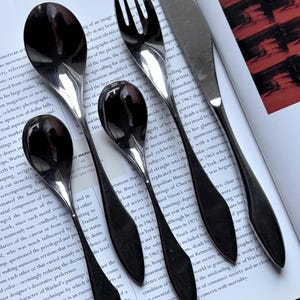 Puede incluir: Un juego de cubiertos plateados pulidos, que incluye cucharas, un tenedor y un cuchillo, dispuestos sobre una página impresa. La cubertería refleja la luz, creando un contraste con el texto impreso. Los utensilios son probablemente para cenar.