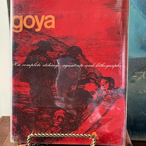 Puede incluir: Un libro rojo titulado "goya" con una ilustración oscura en la portada. El texto "His complete etchings, aquatints and lithographs" es visible. El libro está sobre una pila de otros libros.