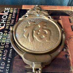Pode incluir: Antigo bate-porta de latão com um design circular com um caractere asiático. O bate-porta tem um design detalhado e ornamentado e está montado em um livro. O texto "CHOICE" é visível.