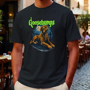Puede incluir: Una camiseta negra de manga corta con un gráfico de un perro marrón con ojos amarillos brillantes tumbado frente a una luna llena y una valla. La palabra Goosebumps está impresa en verde brillante sobre el perro con una fuente goteante.