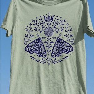 Könnte beinhalten: Ein salbeigrünes Kurzarm-T-Shirt mit einem kreisförmigen dunkelblauen Grafikdruck. Das Design zeigt zwei symmetrische Motten, die sich gegenüberstehen, umgeben von komplizierten Blumen- und Blattmustern in kreisförmiger Anordnung.