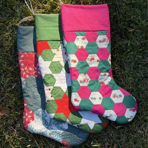 May include: Three quilted Christmas stockings in shades of green, red, and pink, with white hanging loops. The stockings feature geometric patterns, including hexagons and gift box motifs, set against a backdrop of green grass.