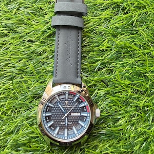 May include: A silver-toned wristwatch with a gray leather band rests on green grass. The watch face has a black geometric pattern with white and red accents. The watch displays the day of the week.