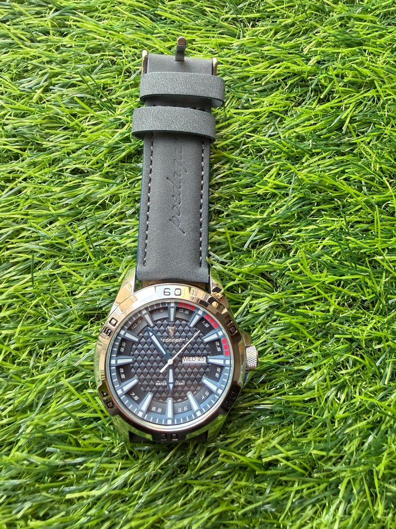May include: A silver-toned wristwatch with a gray leather band rests on green grass. The watch face has a black geometric pattern with white and red accents. The watch displays the day of the week.