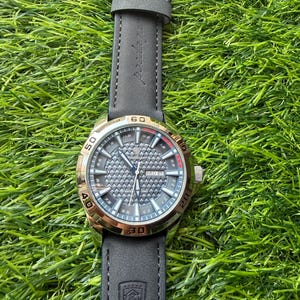 May include: A wristwatch with a gold-colored bezel and a gray dial. The watch has a black leather strap with a buckle. The dial displays the day and date. The watch is set against a background of green grass.