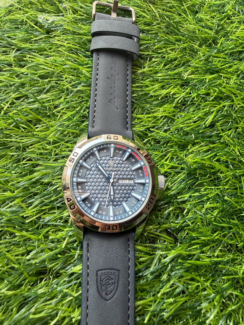 May include: A wristwatch with a gold-colored bezel and a gray dial. The watch has a black leather strap with a buckle. The dial displays the day and date. The watch is set against a background of green grass.