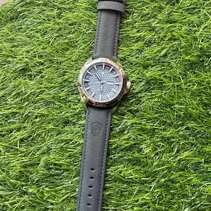 May include: A wristwatch with a black leather band and a gold-colored bezel. The watch face is a textured blue-gray with gold-colored hands and markers. The watch is displayed on a bed of green artificial grass.