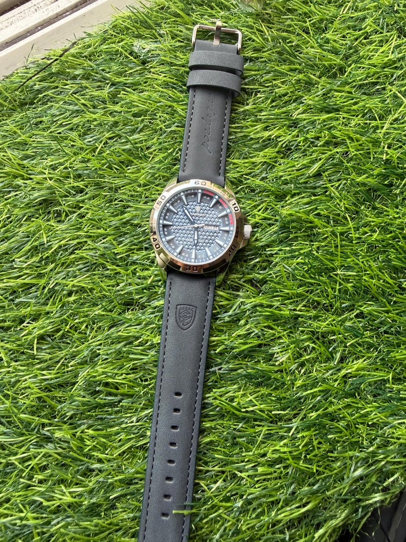 May include: A wristwatch with a black leather band and a gold-colored bezel. The watch face is a textured blue-gray with gold-colored hands and markers. The watch is displayed on a bed of green artificial grass.