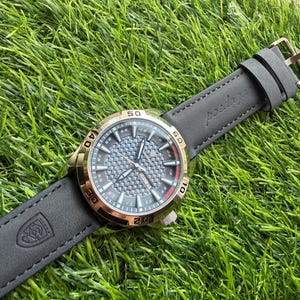 May include: A wristwatch with a gray face and gold-colored bezel, set on a black leather band. The watch face features a textured pattern and visible numerals. The band is embossed with a logo and the word "posdajau".