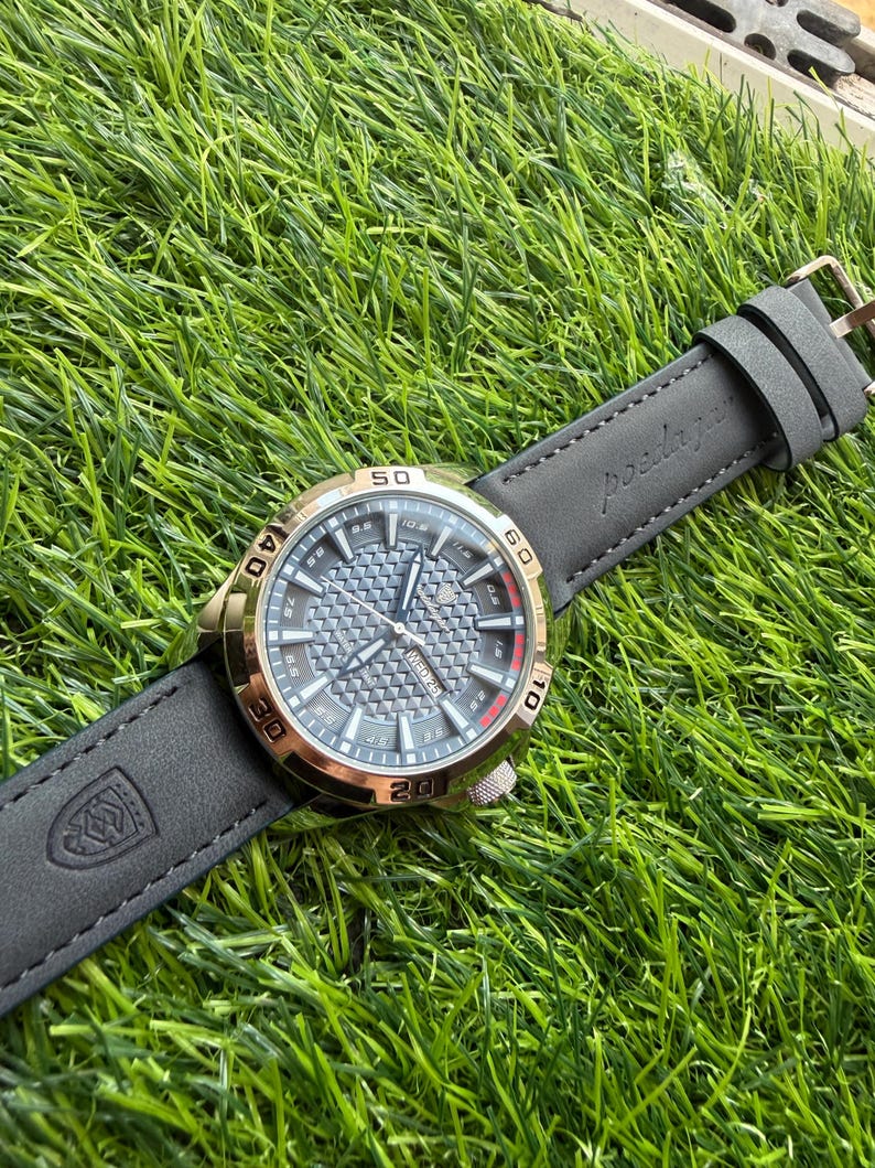 May include: A wristwatch with a gray face and gold-colored bezel, set on a black leather band. The watch face features a textured pattern and visible numerals. The band is embossed with a logo and the word "posdajau".