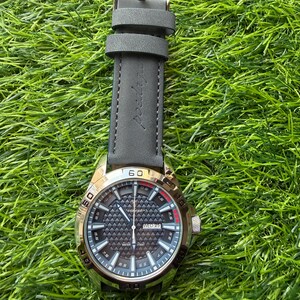 May include: A gold-toned wristwatch with a black patterned face and a gray leather strap. The watch face displays the day and date. The watch is set against a backdrop of green grass.