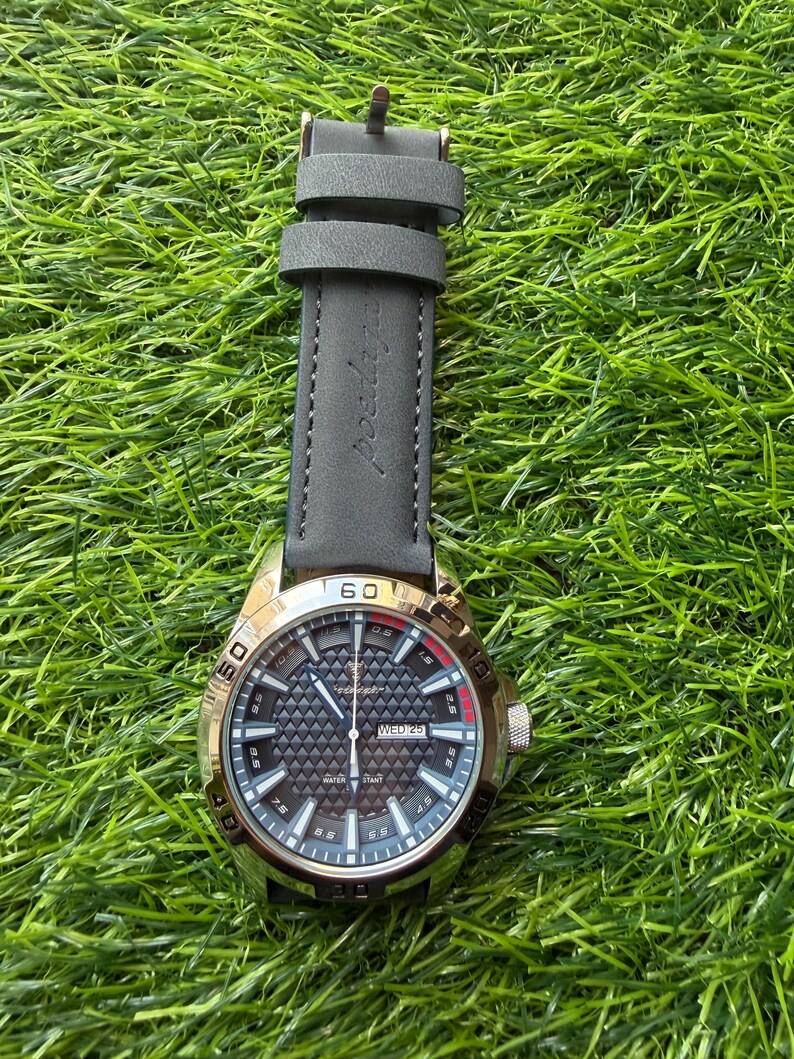 May include: A gold-toned wristwatch with a black patterned face and a gray leather strap. The watch face displays the day and date. The watch is set against a backdrop of green grass.