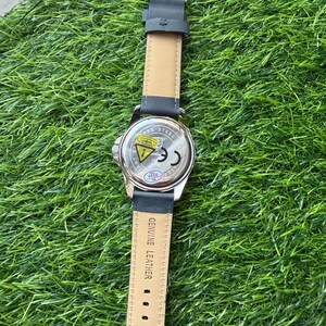May include: A wristwatch with a black leather band and a silver-colored stainless steel case. The watch band is attached with a silver buckle. The back of the watch case has the text "Stainless Steel" and other markings.
