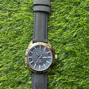 May include: A wristwatch with a black leather band and a gold-colored bezel. The watch face is dark with a textured pattern and features the day and date. The watch is set against a backdrop of green grass.