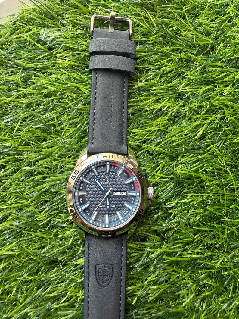 May include: A wristwatch with a black leather band and a gold-colored bezel. The watch face is dark with a textured pattern and features the day and date. The watch is set against a backdrop of green grass.