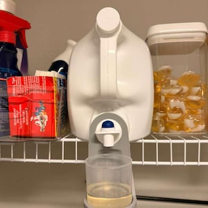 Può includere: Un contenitore bianco di detersivo per bucato con un dispenser attaccato, contenente un liquido trasparente. Il dispenser è grigio con un pulsante blu e un misurino trasparente. Altri prodotti per la pulizia sono visibili sullo scaffale.